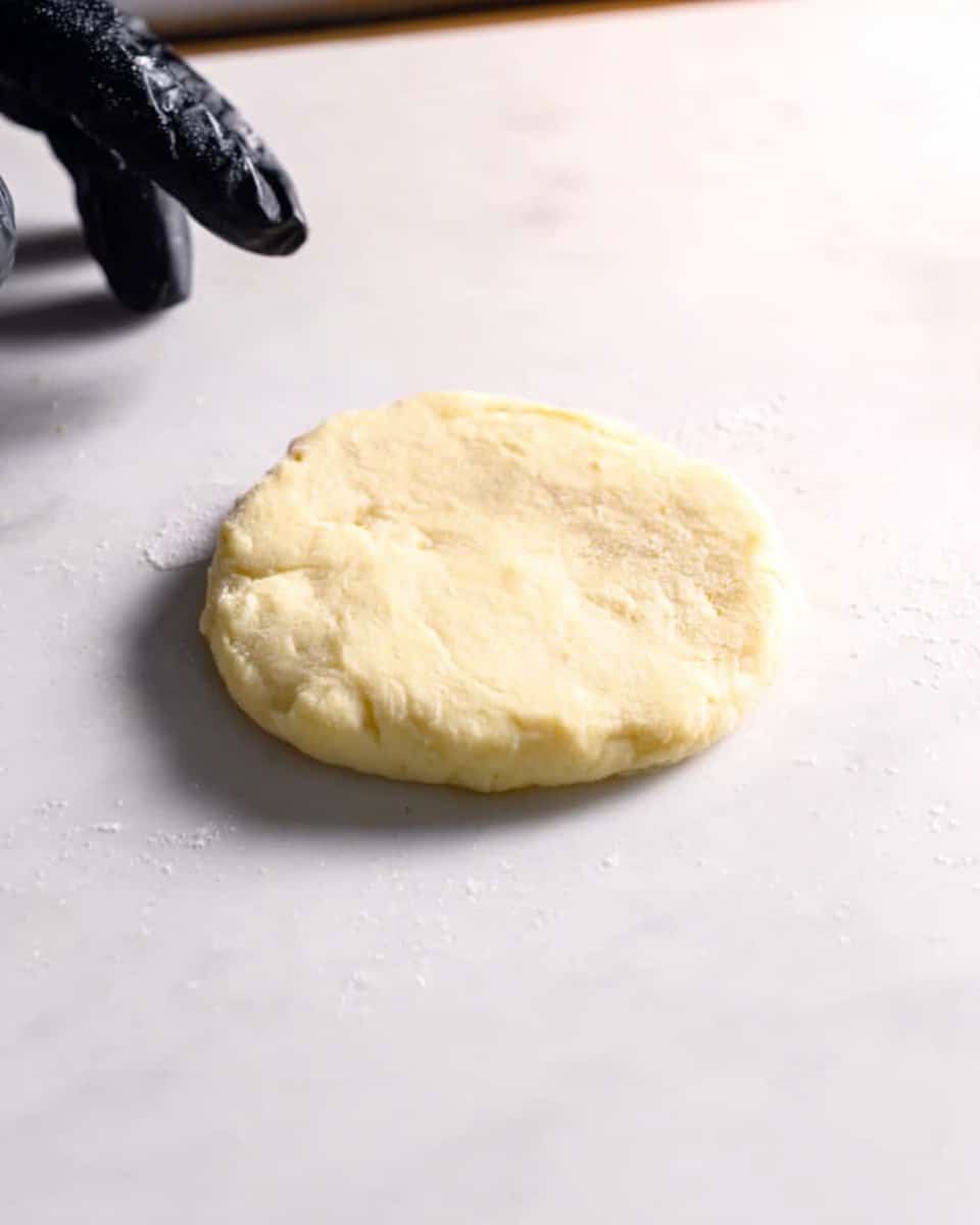 A small, round, flat dough piece of pale yellow color with a slightly rough texture is placed on a white marbled surface. The dough looks soft and smooth with a few uneven edges. A woman's hand wearing a dark glove is seen slightly touching the corner of the dough piece, with no other objects around. The background is simple and clean, highlighting the dough piece in the center. Photo taken with an iphone --ar 4:5 --v 7