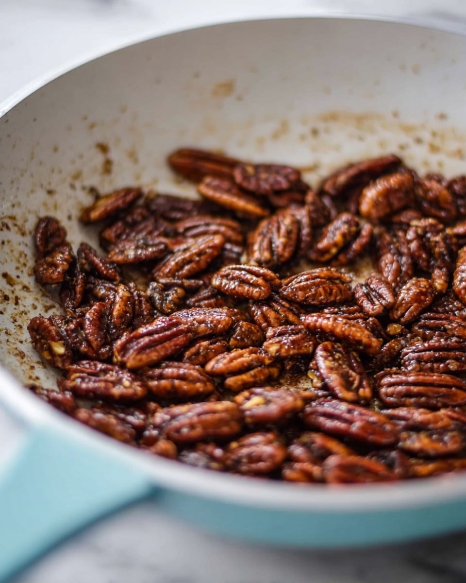 A close-up view of a white pan filled with toasted pecans coated in a glossy brown spice mix. The pecans are in one layer covering the pan's base, showing a rich mix of dark brown and lighter brown tones, with some spice bits scattered around. The pan sits on a white marbled surface, and you can see the pan’s light blue handle coming into view at the bottom left. The image is softly focused on the center pecans, giving a warm and cozy feel. photo taken with an iphone --ar 4:5 --v 7