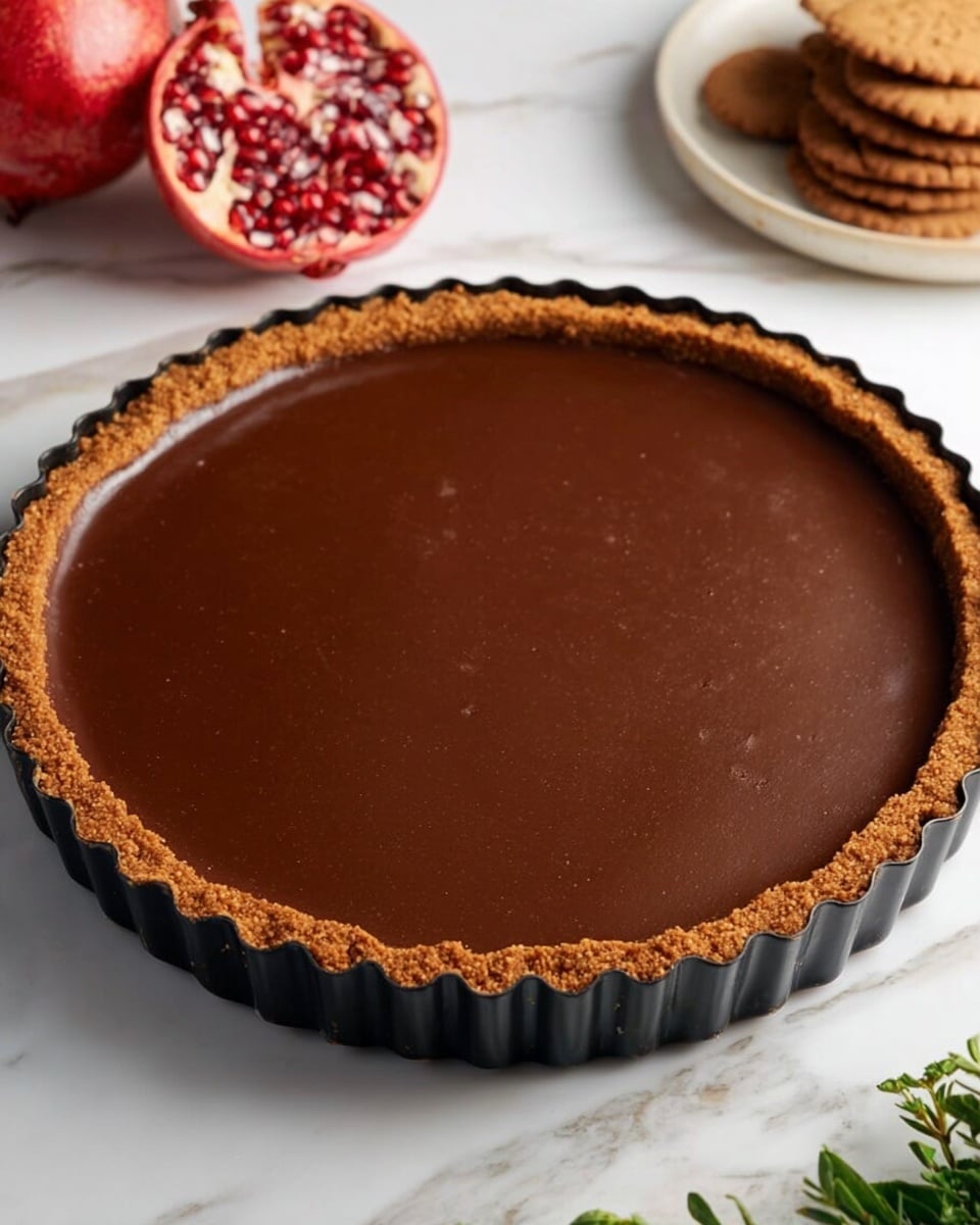 A tart with two visible layers, the bottom layer is a light brown crust forming a thick, uneven edge around the tart, and the top layer is a smooth, thick, dark brown chocolate filling filling the crust completely. The tart sits inside a black fluted tart pan, placed on a white marbled surface. Nearby, there are garnishes showing half a bright red pomegranate with seeds inside and a neat stack of brown gingerbread cookies on the right side. Small green leaves are visible at the bottom right corner, adding contrast to the scene. Photo taken with an iphone --ar 4:5 --v 7