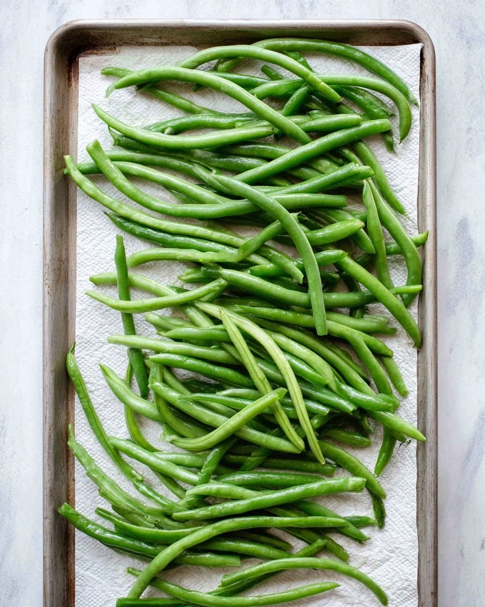 A single layer of fresh, green beans spread out evenly on a metal baking tray lined with white paper towels, the green beans are bright and shiny with smooth textures, their long, slender shapes slightly curved and overlapping one another, placed on a white marbled surface photo taken with an iphone --ar 4:5 --v 7