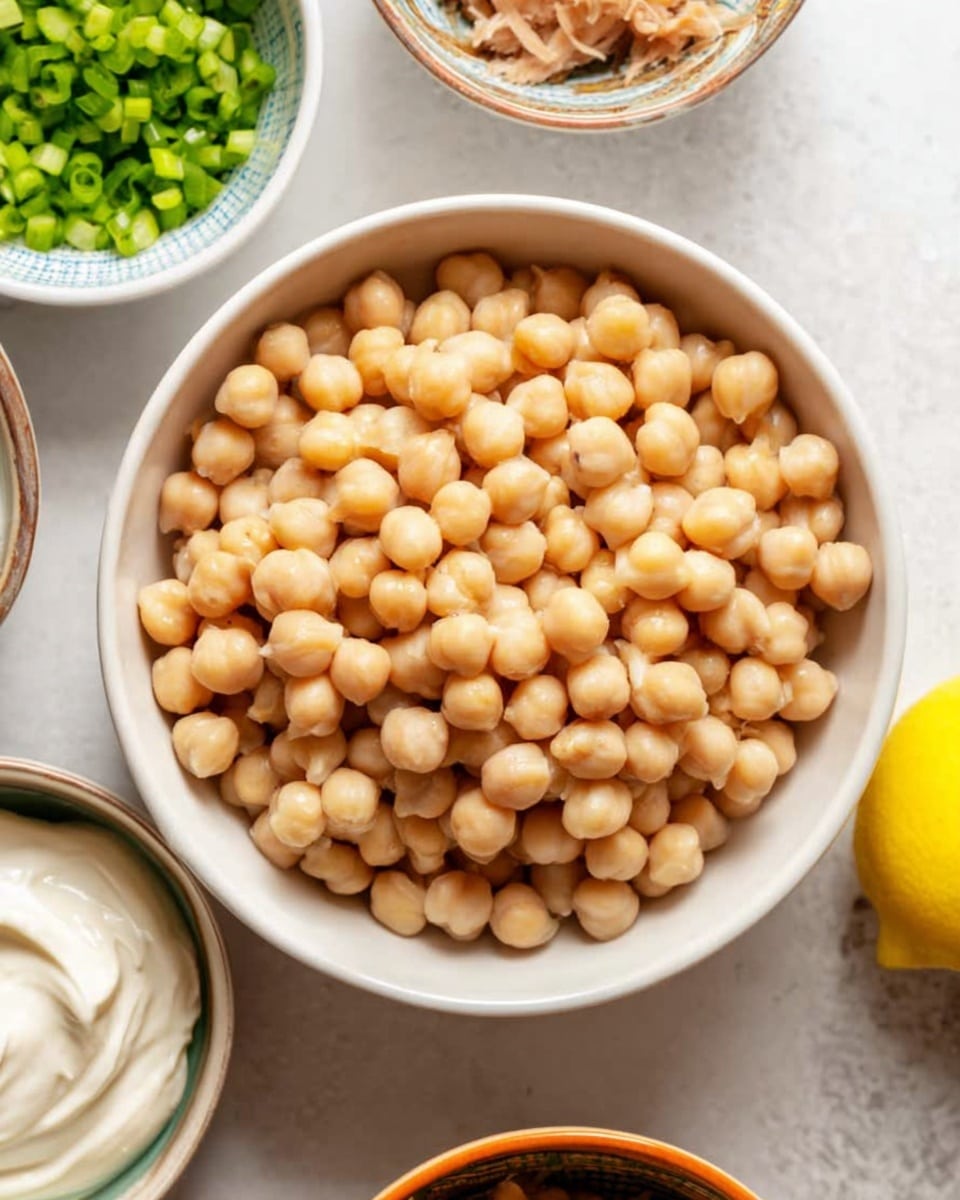 The image shows two white bowls on a white marbled surface; the first bowl contains a creamy yellow sauce being stirred with a metal whisk, the texture smooth with small black specks throughout, coating the sides of the bowl. The second bowl is filled with mixed layers: the base is light beige chickpeas, topped unevenly with chopped green celery, dark green herbs, finely diced purple onions, small capers, and shredded light brown tuna, all covered in the same creamy yellow sauce drizzled over the top, creating a rich, layered look. photo taken with an iphone --ar 4:5 --v 7