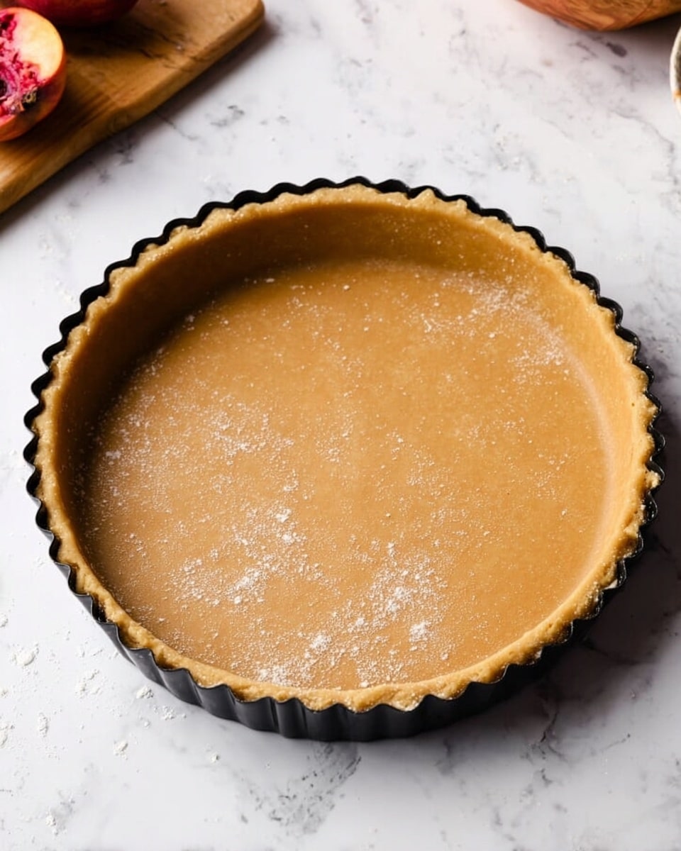 A round tart pan filled with smooth, even light brown dough pressed flat into the base and up the sides, creating a single uniform layer with a slightly crimped edge along the pan's rim, sitting on a white marbled surface. There are faint traces of flour dusted lightly over the dough’s surface. The pan is black with a scalloped edge. In the background, a wooden cutting board with a halved red fruit is visible at the top left corner. Photo taken with an iphone --ar 4:5 --v 7