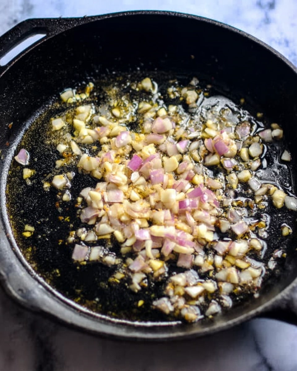 A black cast iron pan filled with small, light yellow chopped garlic and light purple chopped onions frying in clear oil. The garlic and onions are spread evenly in a single thin layer across the pan, showing a mix of soft and slightly shiny textures. The pan rests on a white marbled surface. photo taken with an iphone --ar 4:5 --v 7