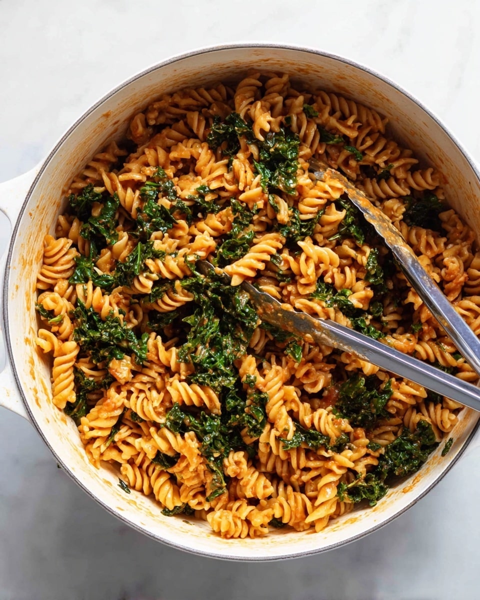 A white pot filled with twisted spiral pasta cooked with dark green leafy kale, all coated in a light reddish-orange sauce. The pasta spirals are evenly mixed with the kale, showing the rough leaf texture against the smooth pasta. A metal fork and tongs rest inside the pot, partly mixing the contents. The pot sits on a white marbled surface. photo taken with an iphone --ar 4:5 --v 7