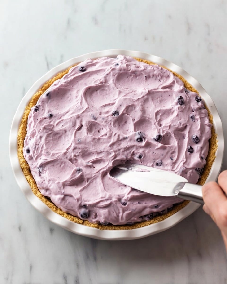 The image shows a round pie with three layers. The bottom layer is a golden brown crust, firm and even, filling a white pie plate with a scalloped edge. On top of the crust is a thick, fluffy layer of light purple cream filling that looks smooth and dotted with small dark berry pieces throughout. A woman's hand is holding a spatula, spreading the cream evenly on the pie’s surface but leaving soft peaks and swirls in the creamy texture. The pie sits on a white marbled surface. photo taken with an iphone --ar 4:5 --v 7