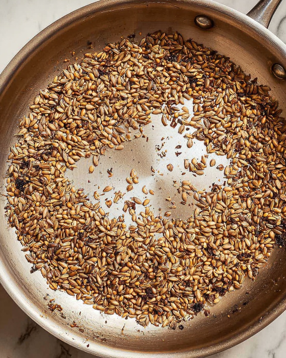 A close-up view of a silver metal pan with a single layer of small, elongated seeds being toasted. The seeds range in color from light beige to dark golden brown, with some showing small blackened spots, indicating uneven toasting. The center of the pan shows the smooth, shiny metal surface with no seeds, while the seeds spread evenly around the edges, creating a rough ring shape. The pan has a reflective surface with slight discoloration from cooking. The background is a white marbled texture. photo taken with an iphone --ar 4:5 --v 7