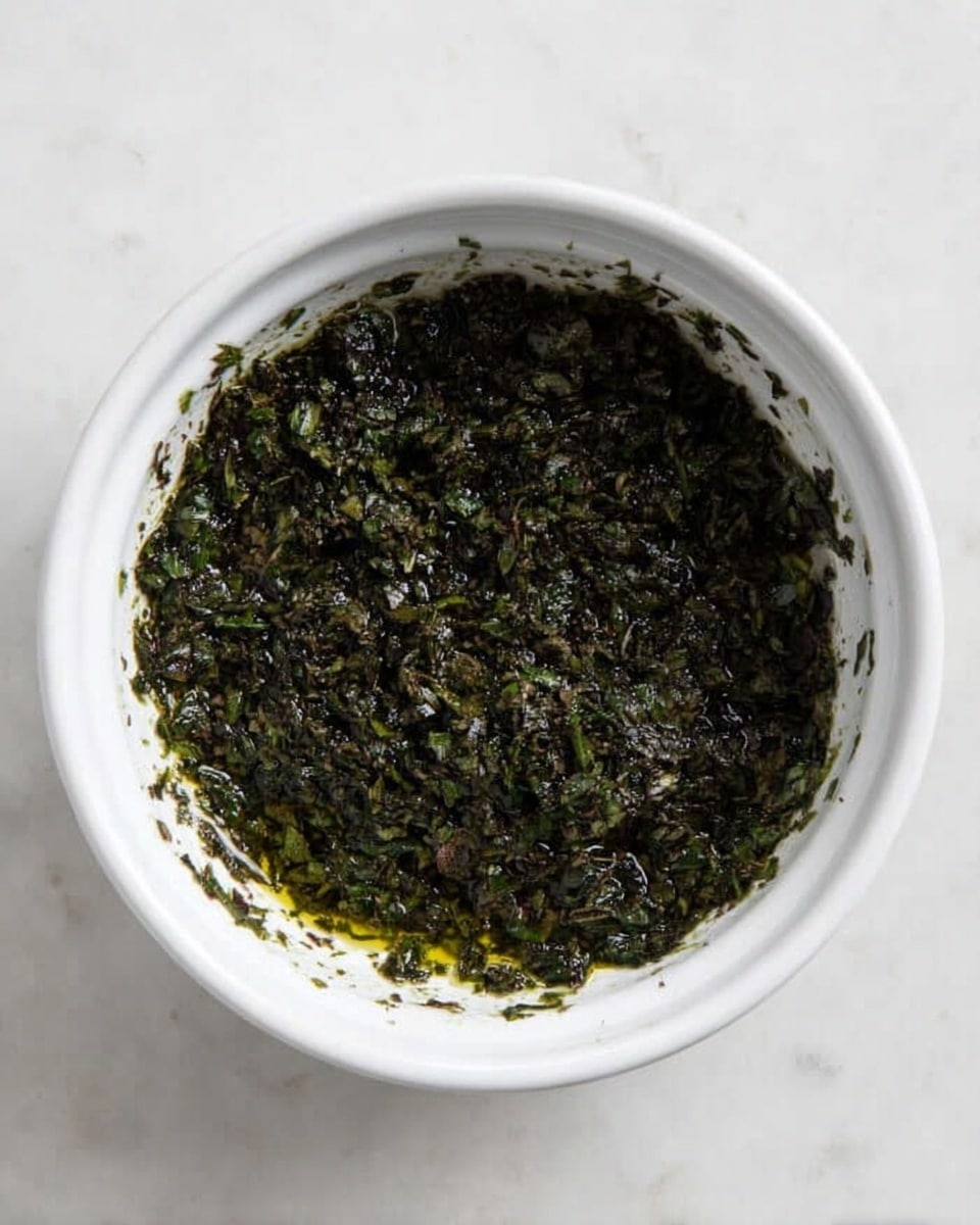 A white bowl filled with a dark green, finely chopped herb mixture soaked in oil. The mixture covers the bottom and edges of the bowl with a slightly wet texture, showing small bits of leaves and stems throughout. The bowl sits on a white marbled surface. photo taken with an iphone --ar 4:5 --v 7