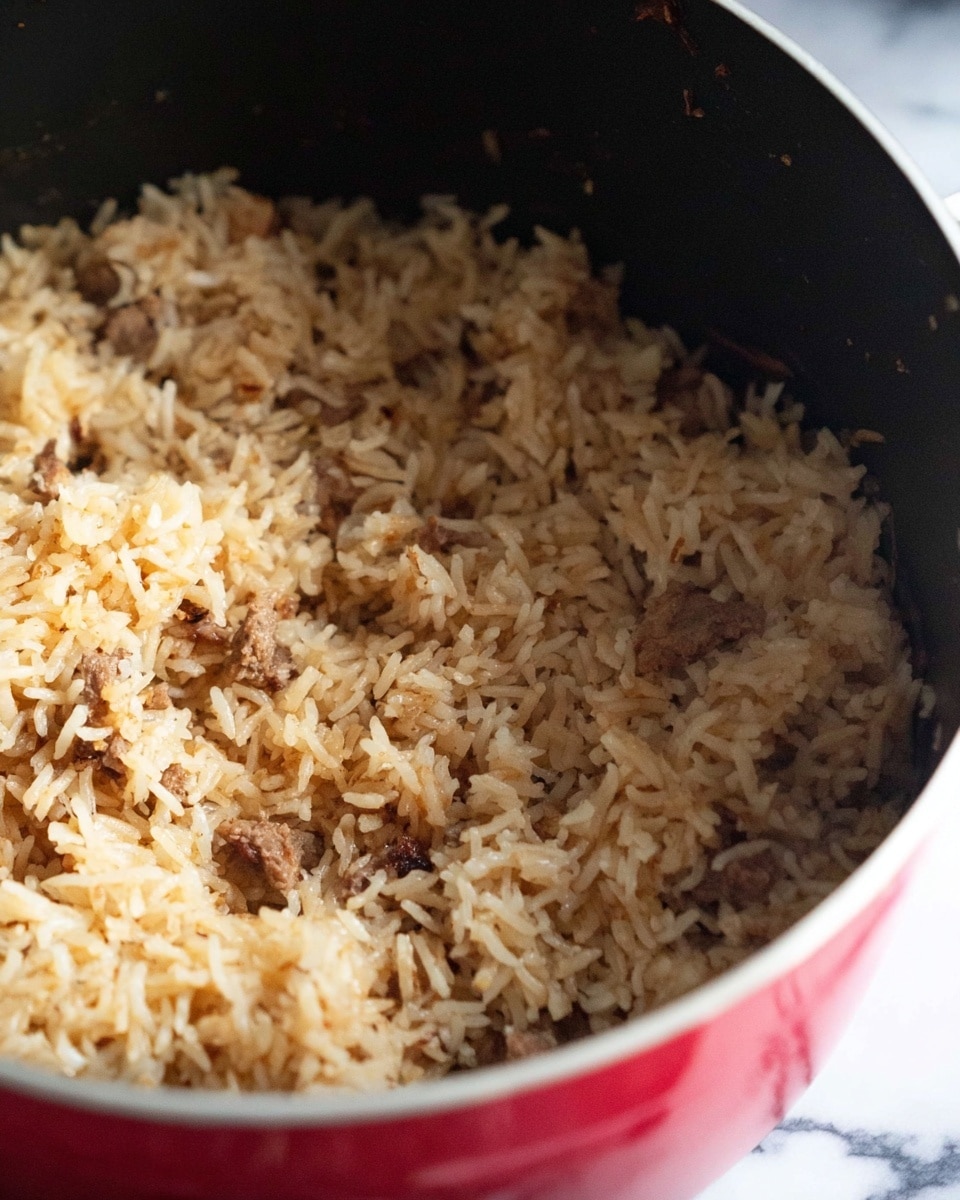 The image shows a pot filled with cooked rice mixed with small pieces of meat. The rice is light brown with a fluffy texture, and the meat is scattered evenly throughout, appearing darker brown with a slightly crumbly look. The pot is red on the outside with a black interior, sitting on a white marbled surface. The focus is on the rice and meat combination inside the pot, showing the grain details and bits of meat closely. photo taken with an iphone --ar 4:5 --v 7
