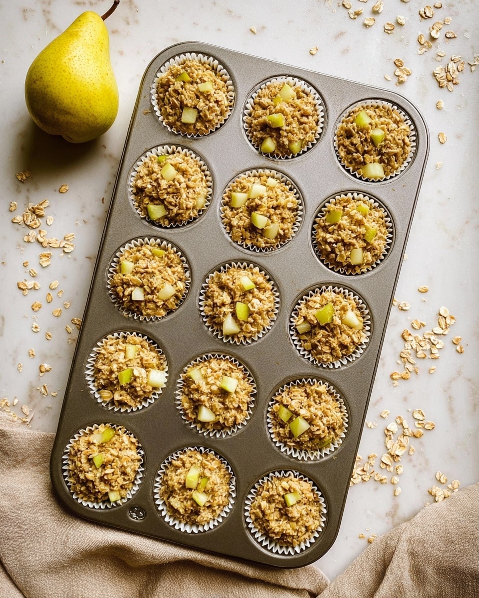 The image shows a metal muffin tray with twelve cups, each filled with a mixture of oats and small greenish-yellow pear pieces. The oats look soft and slightly wet, creating a crumbly texture. Pear chunks are scattered evenly in each cup, giving a mix of light green and pale yellow colors. The tray rests on a white marbled surface with some oats and pear bits scattered around. A whole pear with a yellow-green skin is placed to the left of the tray, and a beige cloth is partially visible on the bottom right corner. Photo taken with an iphone --ar 4:5 --v 7