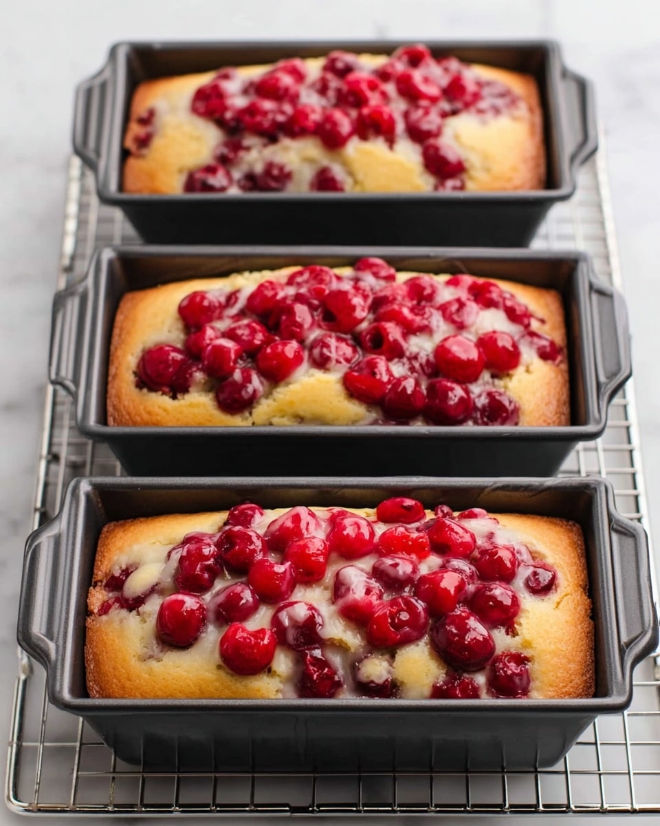 Three small rectangular black baking pans sit side by side on a metal cooling rack over a white marbled surface. Each pan holds a loaf of light golden-yellow cake, topped with a generous layer of bright red cherries that have a glossy texture and are slightly sunken into the soft cake. The cake layer is smooth and slightly risen in the middle, with some cracks around the cherry topping showing the moist texture inside. photo taken with an iphone --ar 4:5 --v 7