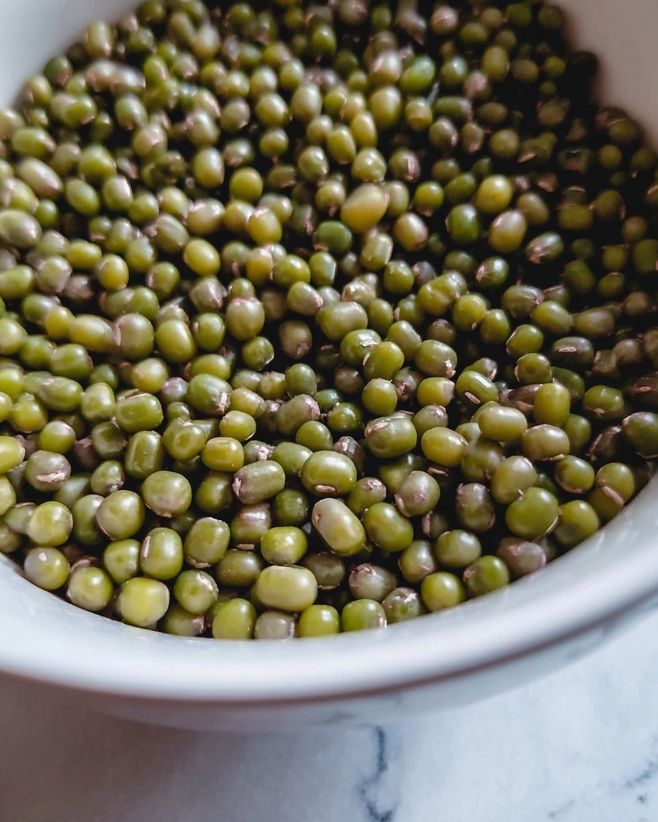 A close-up view of a white bowl filled with a large pile of small green mung beans. The mung beans are smooth, round, and vary slightly in shades of green and light brown, tightly packed inside the bowl. The bowl's inner surface is shiny and white, reflecting some light, and the background has a white marbled texture. The image focuses on the beans with a slight shallow depth of field, showing clear texture and details on the mung beans' surface. photo taken with an iphone --ar 4:5 --v 7