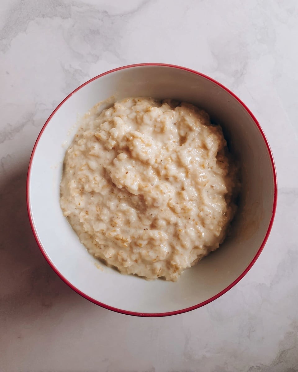 A white bowl with a red rim contains a creamy oatmeal base topped with three fresh red strawberries with green leaves arranged in a line on one side, a cluster of five dark blackberries below the strawberries, thin light beige almond flakes placed next to the strawberries on the left, and a drizzle of amber-colored syrup over the oatmeal in the center. The bowl is set on a white marbled surface scattered with oatmeal flakes, next to a gray textured cloth and a silver spoon. photo taken with an iphone --ar 4:5 --v 7