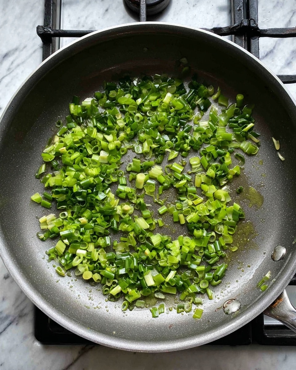 A frying pan filled with small, chopped green scallions spread evenly across the surface. The scallions show different shades of green, from light to dark, and some small drops of oil glisten around them. The pan is set on a stove burner, with the knob visible on the side. The background is a white marbled texture. photo taken with an iphone --ar 4:5 --v 7