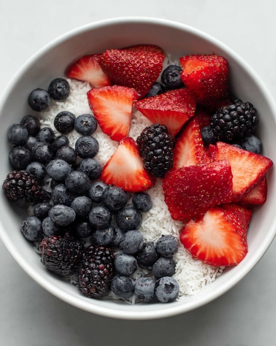 A white bowl filled with three layers of fresh fruit, starting with a bottom layer of lightly textured white coconut flakes, followed by a middle layer of small, round dark blue blueberries scattered evenly, and topped with a mix of bright red strawberry slices and shiny blackberries spread throughout the bowl. The strawberries show a vibrant red color with white centers and small seeds, adding a fresh look to the mix. The bowl sits on a white marbled surface. photo taken with an iphone --ar 4:5 --v 7