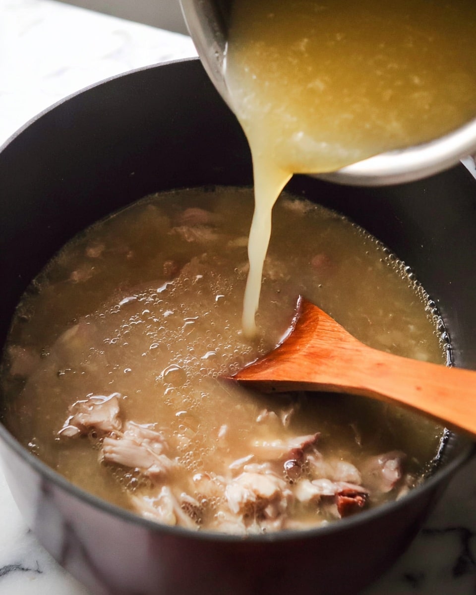 A close-up view of a dark-colored pot filled with a thick, light brown soup containing chunks of white meat and bits of darker meat mixed in. A wooden spoon with a smooth surface is partially submerged in the soup, stirring gently. Above the pot, a silver container pours a light yellowish broth into the pot, adding more liquid. The image shows bubbles on the soup surface, indicating it is hot and simmering. The background is a white marbled texture. photo taken with an iphone --ar 4:5 --v 7