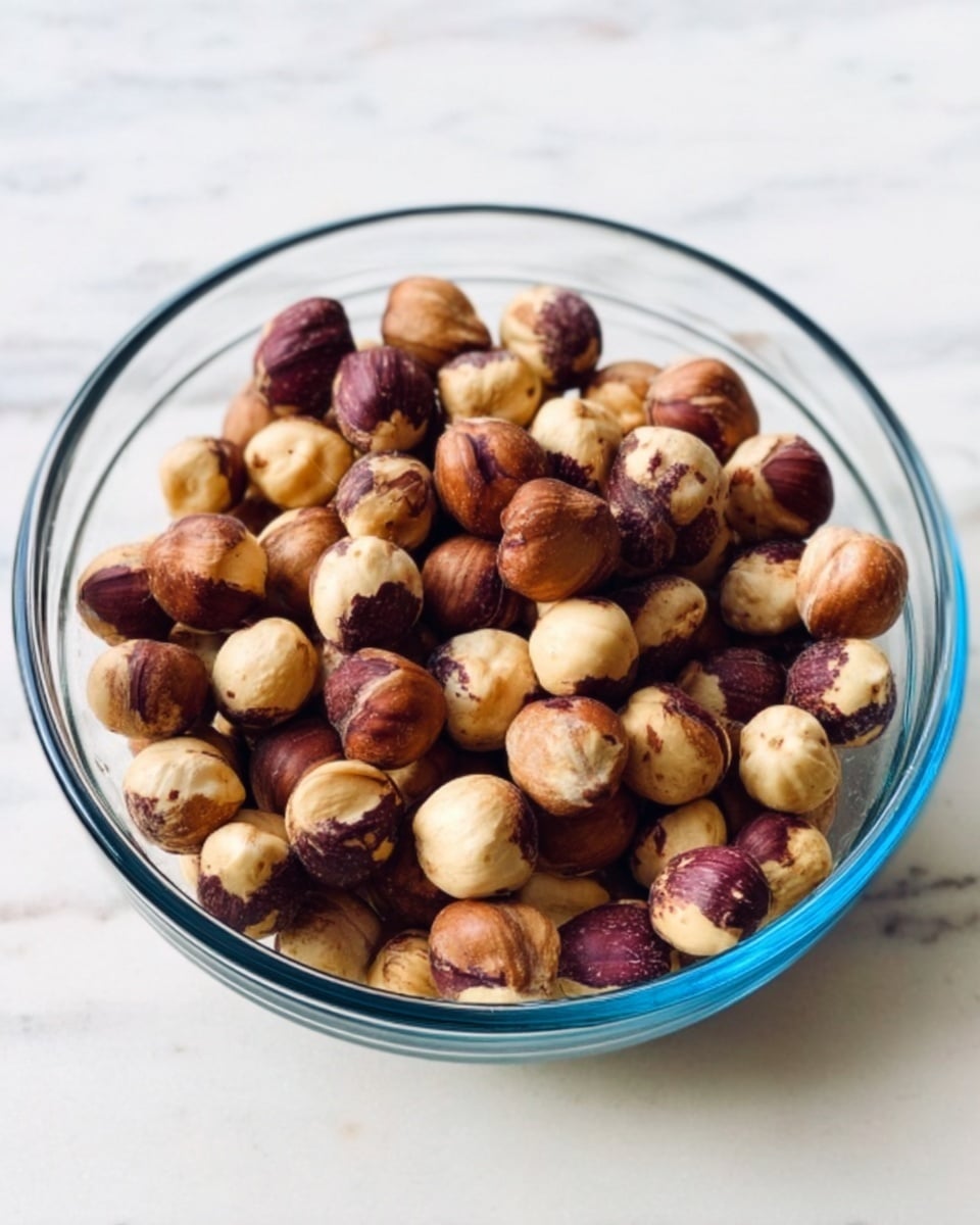 A clear glass bowl filled with light and dark brown roasted hazelnuts, showing a smooth, slightly shiny texture. The bowl is placed on a white marbled surface, with natural light highlighting the round nuts and their varied colors. The hazelnuts fill the bowl evenly, some showing a split shell texture. Photo taken with an iphone --ar 4:5 --v 7