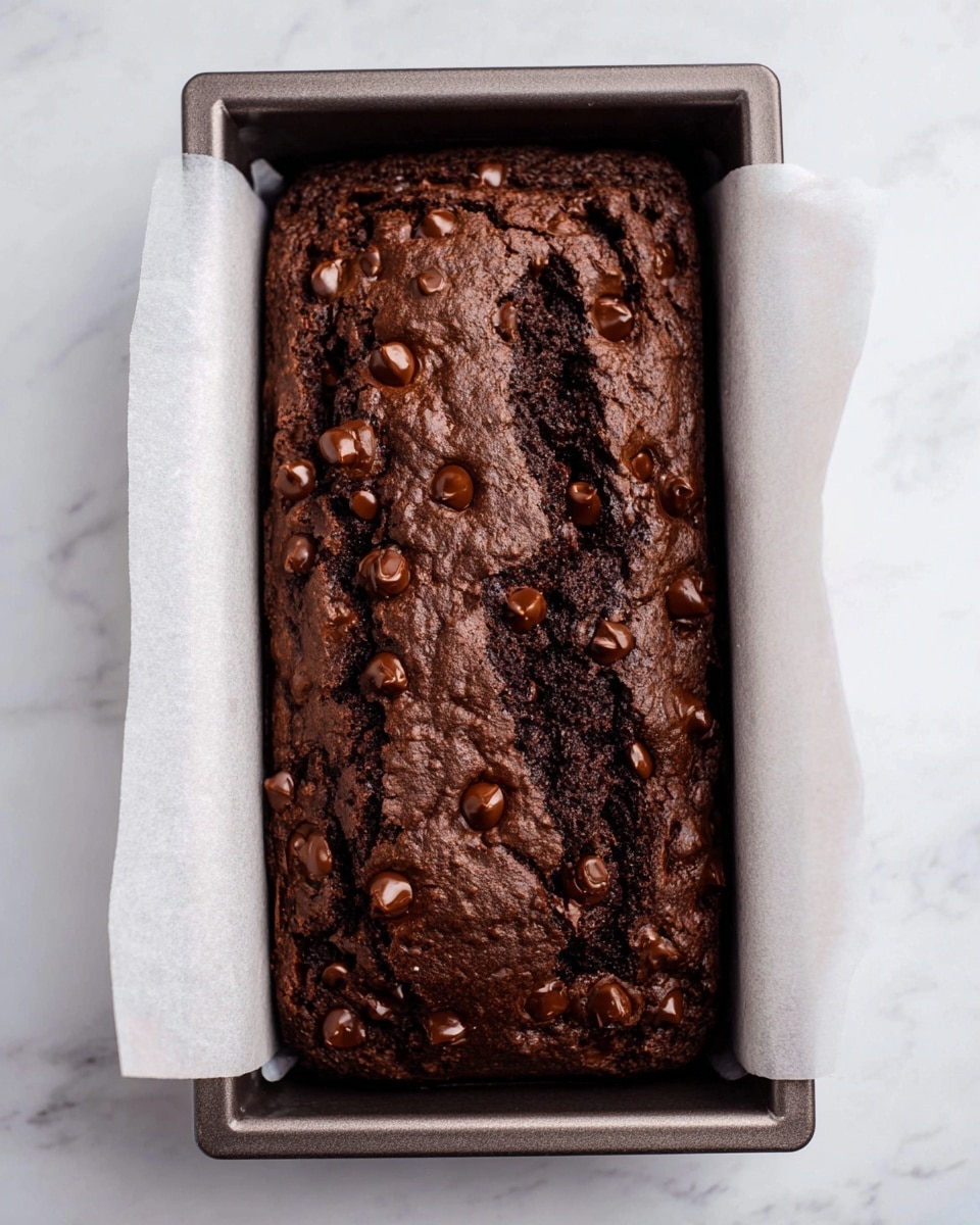 A freshly baked dark brown chocolate cake with a cracked top is shown inside a rectangular metal baking tray lined with white baking paper. The cake's surface is sprinkled with many small, shiny chocolate chips that add texture. The cake looks soft with a rough and slightly bumpy surface. The tray sits on a white marbled surface. photo taken with an iphone --ar 4:5 --v 7