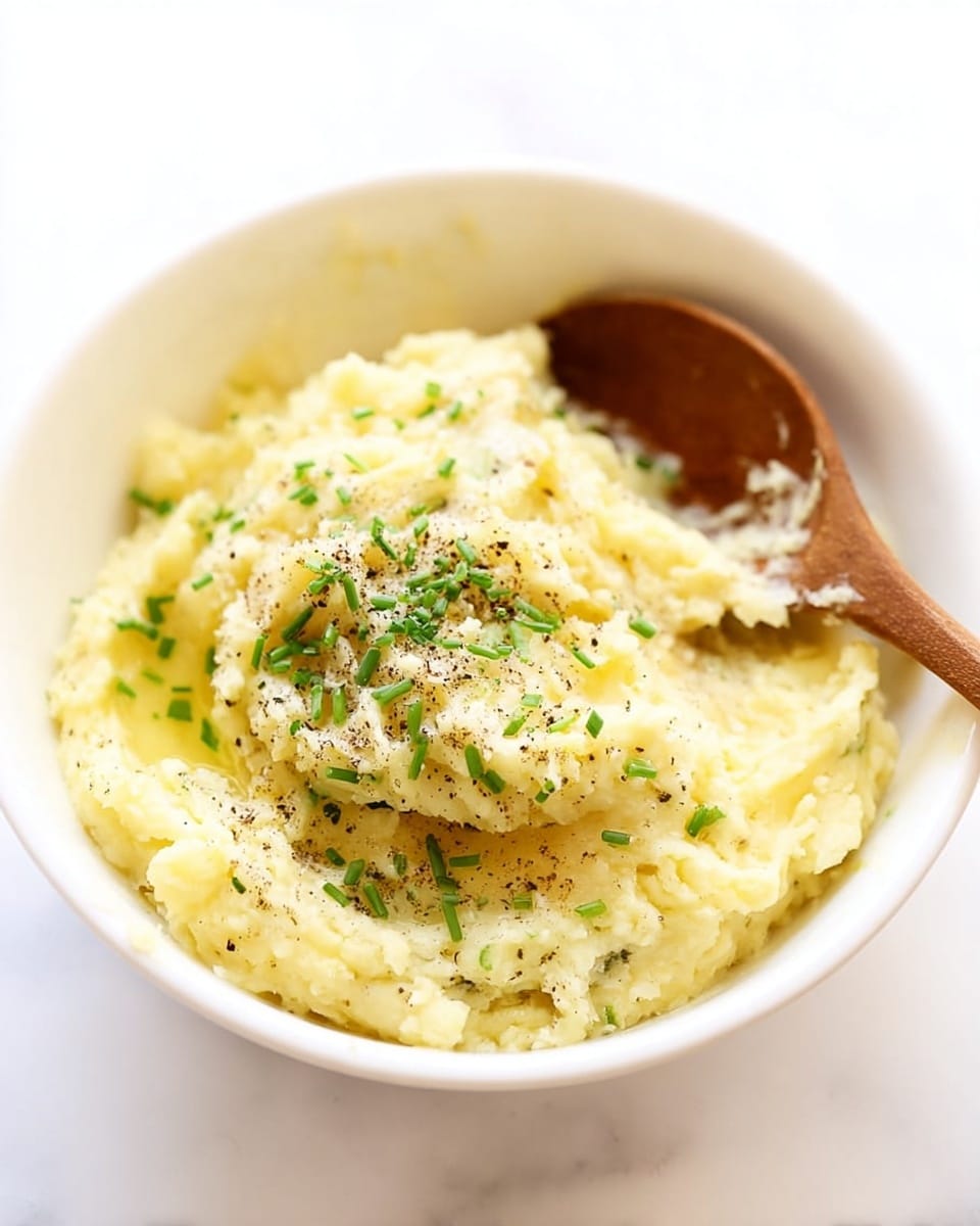 The dish shows a round bowl filled with creamy mashed potatoes that have a light yellow tone with some small chunks for texture. On top, there is a square piece of melting butter with tiny bits of chopped green chives sprinkled all over. The bowl is placed on a white marbled surface, with some whole chives, a garlic clove, and an onion faintly blurred in the background. photo taken with an iphone --ar 4:5 --v 7