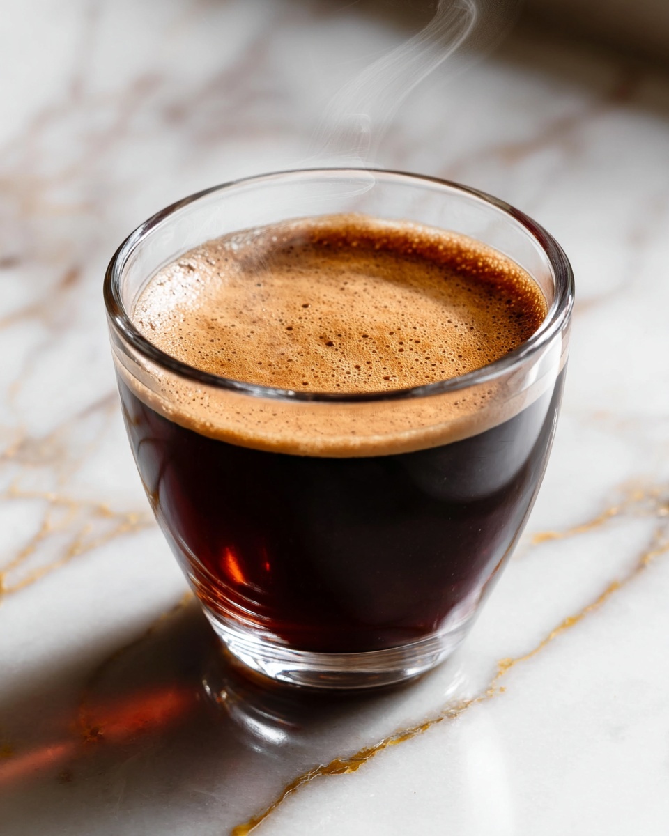 A clear glass cup filled with dark brown coffee sits on a white marbled surface with gold veins. The coffee has a smooth, light brown froth layer on top about one centimeter thick, with a few small bubbles and an even texture. Thin steam rises gently from the coffee, creating soft curves above the cup. The shiny glass catches light, showing reflections and the dark coffee’s rich color inside. The white marbled surface underneath has subtle gray lines and looks clean and polished. Photo taken with an iphone --ar 4:5 --v 7