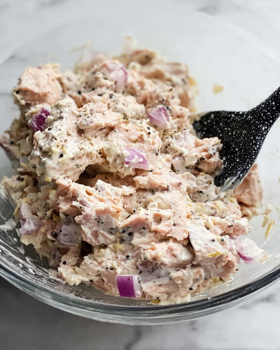 A clear bowl contains a creamy mixture with small chunks of light pink tuna and bits of purple onion scattered throughout. The texture looks soft and slightly fluffy with some small black pepper specks visible. A black spatula with white speckled pattern is partially submerged in the mixture, resting inside the bowl. The background shows a white marbled surface. photo taken with an iphone --ar 4:5 --v 7