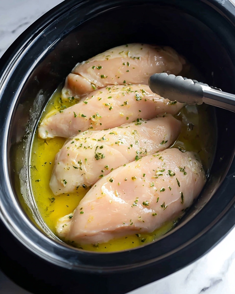 A black slow cooker filled with three raw chicken breasts laying side by side. The chicken is light pink with a smooth, moist texture, and small green herb pieces sprinkled on top. A light yellow marinade with a slightly oily surface surrounds the chicken, pooling around the edges. Stainless steel tongs grip one piece of chicken on the right side. The background shows a white marbled surface. Photo taken with an iphone --ar 4:5 --v 7