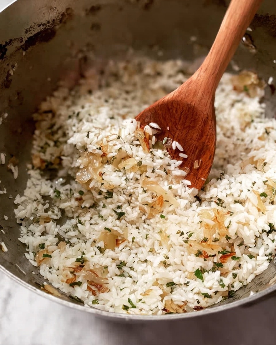 A close-up image shows a pot with short white grains of rice mixed with small pieces of cooked light brown onion and green herbs. The rice grains have a slightly shiny, soft texture. A wooden spoon is stirring the rice mixture, with part of the spoon's bowl lifting some rice. The inside of the pot has a metallic surface, reflecting light and showing some cooked bits stuck to it. The background surface is a white marbled texture. photo taken with an iphone --ar 4:5 --v 7