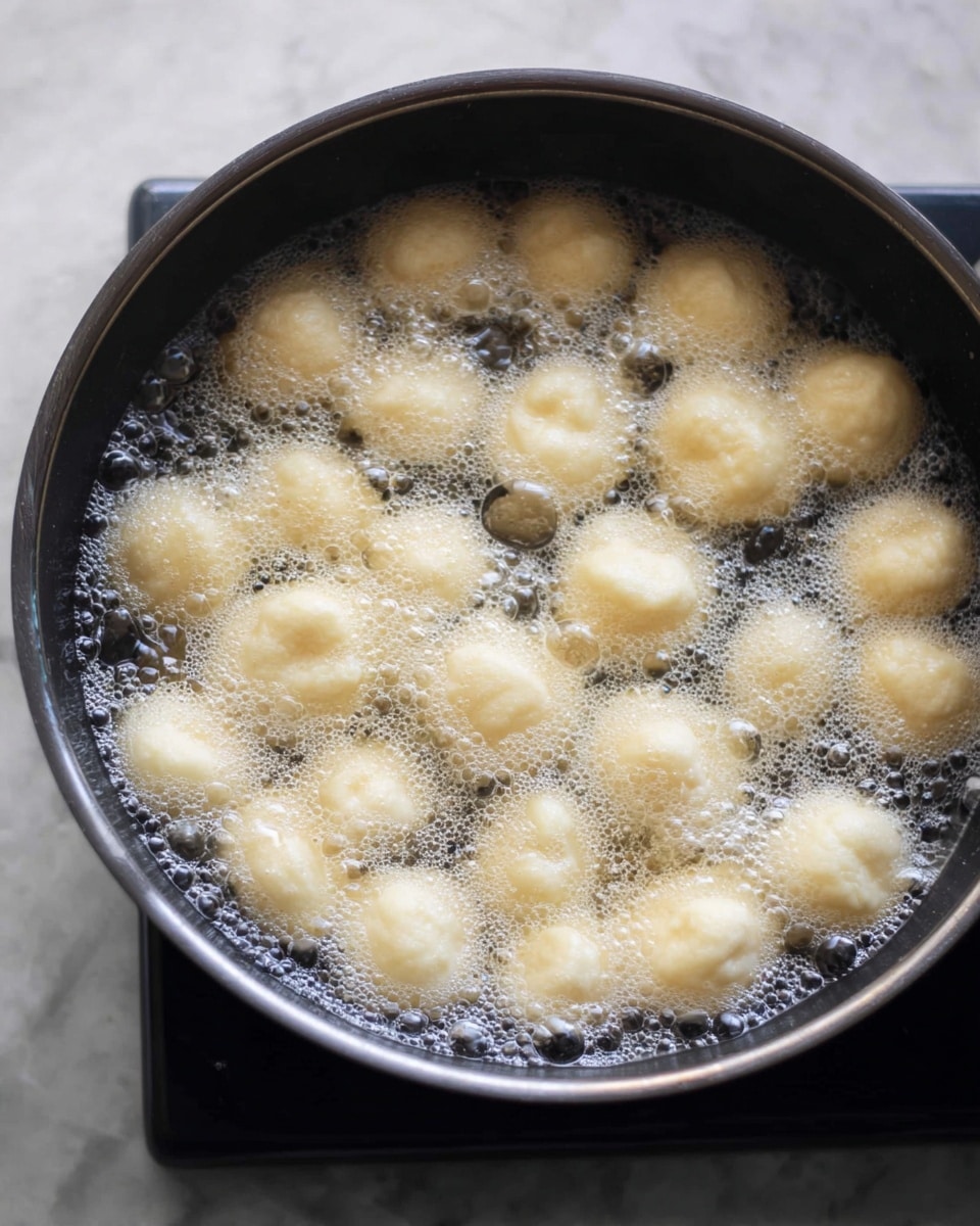 A black pot filled with clear boiling water showing many small, pale yellow dumplings floating and cooking on the surface, surrounded by lots of small bubbles around each dumpling, creating a soft foam on top, all placed on a white marbled surface photo taken with an iphone --ar 4:5 --v 7