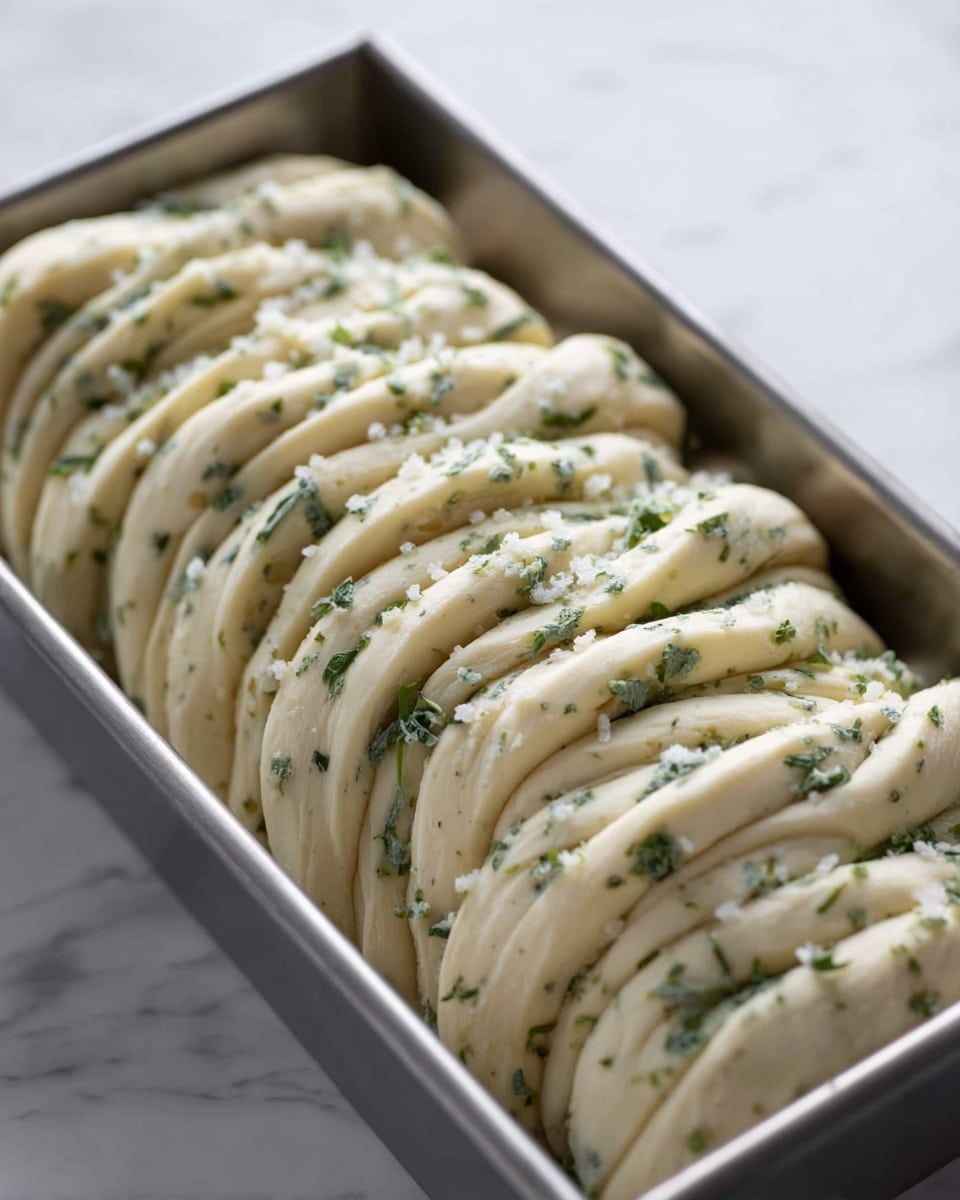 A close-up view of a rectangular metal baking pan filled with many layers of light beige dough, each layer thick and soft with green herb bits spread between them. The dough layers are folded and placed closely together, creating a repetitive wave-like structure. Small grains of white salt or sugar crystals are sprinkled on top, adding a slight texture contrast. The pan sits on a white marbled surface, giving a clean and fresh background. photo taken with an iphone --ar 4:5 --v 7
