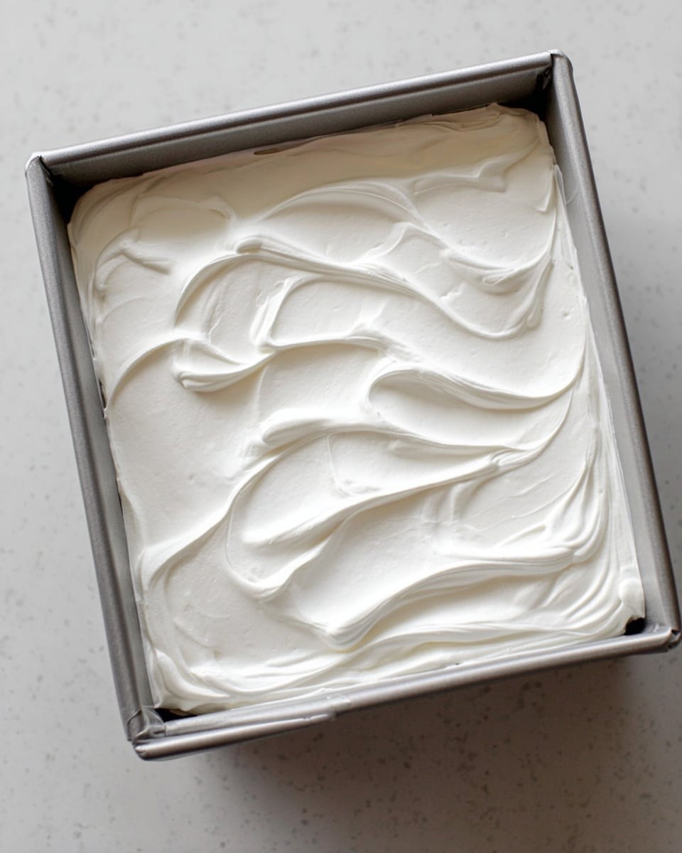 A silver square baking pan filled with one thick layer of smooth, white whipped cream or frosting, spread evenly with soft, wavy swirls on the surface. The pan is placed on a white marbled countertop, providing a clean and bright background. photo taken with an iphone --ar 4:5 --v 7