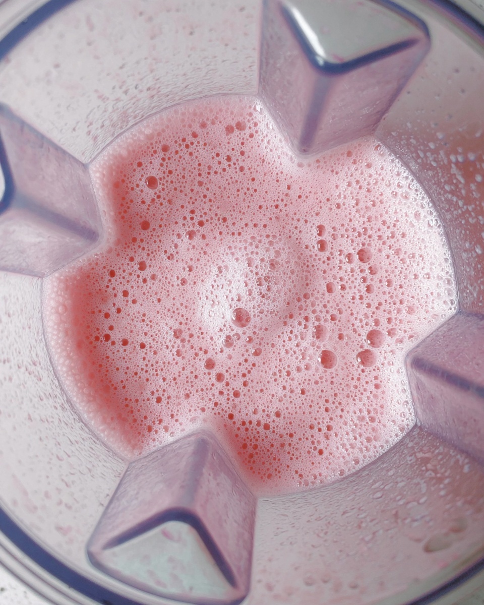 A top view of a blender filled with a layer of light pink frothy liquid that has many small bubbles over the surface. The blender is clear, showing the thick, smooth texture of the pink mix inside. The background is a white marbled texture. photo taken with an iphone --ar 4:5 --v 7