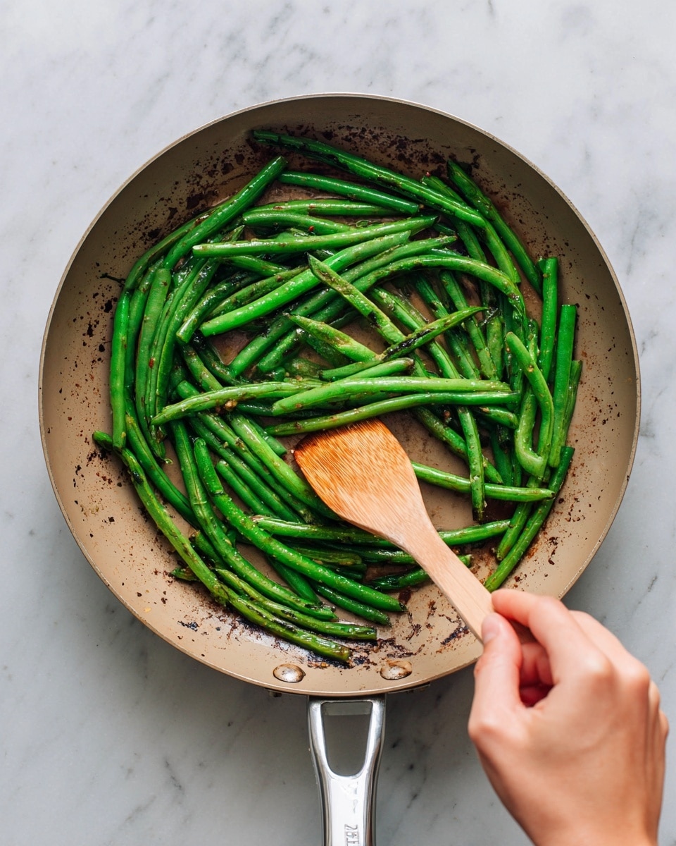 A round metal pan filled with bright green cooked green beans scattered with small toasted pale brown almond slivers and bits of garlic, with some slight charring on the beans and pan edges. A wooden spoon with a warm brown color rests inside the pan, held by a woman's hand at the bottom of the image stirring the beans. The pan is placed on a white marbled surface that adds a clean, neutral background to the scene. photo taken with an iphone --ar 4:5 --v 7