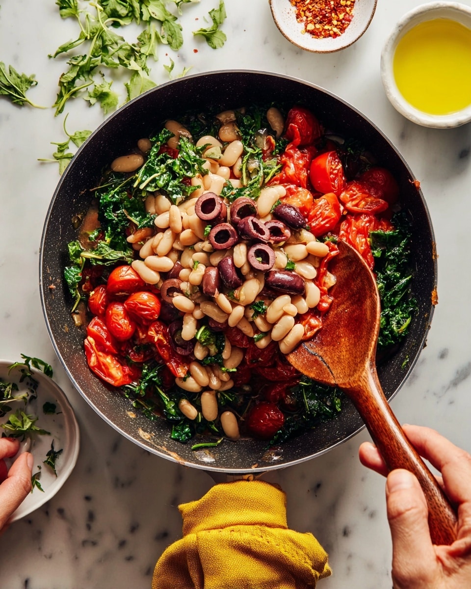 A black pan sits on a white marbled surface filled with a dish that has multiple layers: the bottom layer is soft cooked red cherry tomatoes with a shiny, slightly wrinkled texture; above this is a bed of chopped dark green leafy vegetables spread unevenly; on top is a mix of light beige white beans and sliced dark purple olives scattered across the greens and tomatoes. A woman's hand holding a wooden spoon with a rich brown color is stirring the contents from the left side, while another woman's hand grips the pan handle wrapped with a mustard yellow cloth on the bottom right. Nearby, there are some scattered leafy greens, a white spoon with red chili flakes, and a white bowl with a yellow liquid, all on the white marbled surface. Photo taken with an iphone --ar 4:5 --v 7