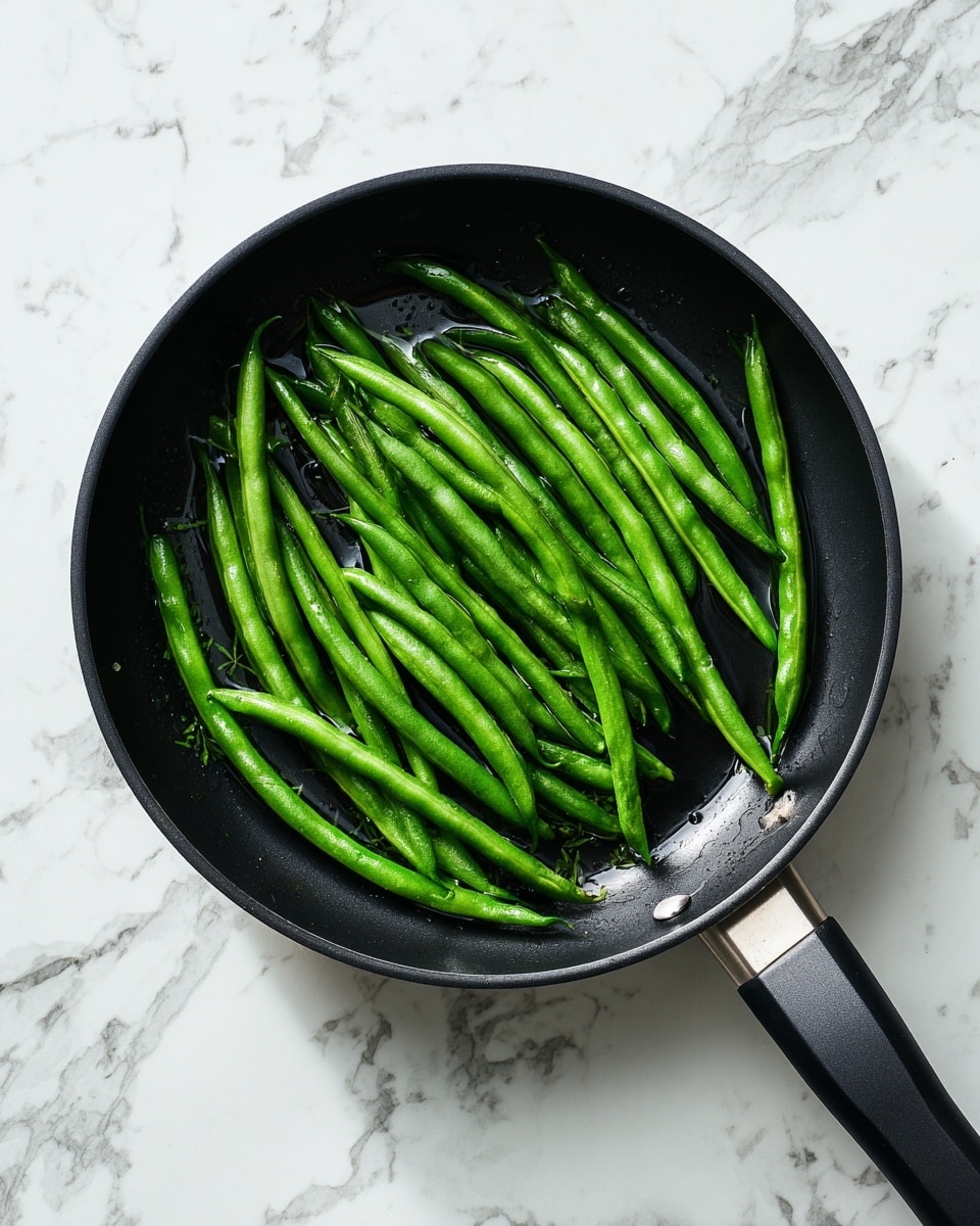 A black pan with a long black handle contains a single layer of bright green long beans. The beans are fresh and glossy, arranged loosely and evenly across the pan’s surface with some water visible underneath them. The pan rests on a white marbled surface, showing a clean and simple kitchen setting. Photo taken with an iphone --ar 4:5 --v 7