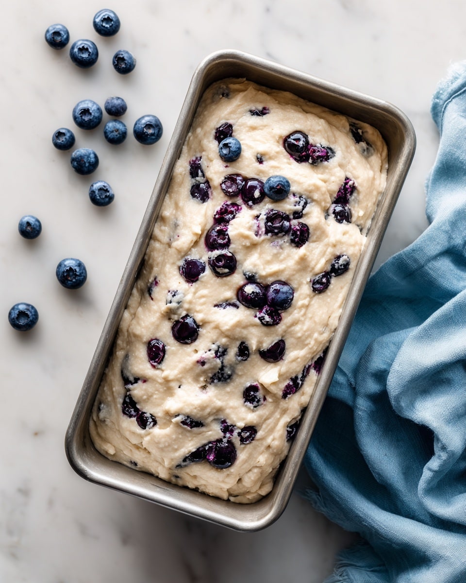 The image shows a silver rectangular baking pan filled with thick light beige batter that has a slightly rough texture. Scattered throughout the batter are whole blueberries, some embedded inside and some sitting on top, adding dark blue and purple spots to the surface. The pan is placed on a white marbled surface with a few loose blueberries nearby on the left. On the right side of the pan, there is a soft, light blue cloth casually draped. The scene is softly lit, and the image is taken from above, giving a clear view of the batter and blueberries. photo taken with an iphone --ar 4:5 --v 7