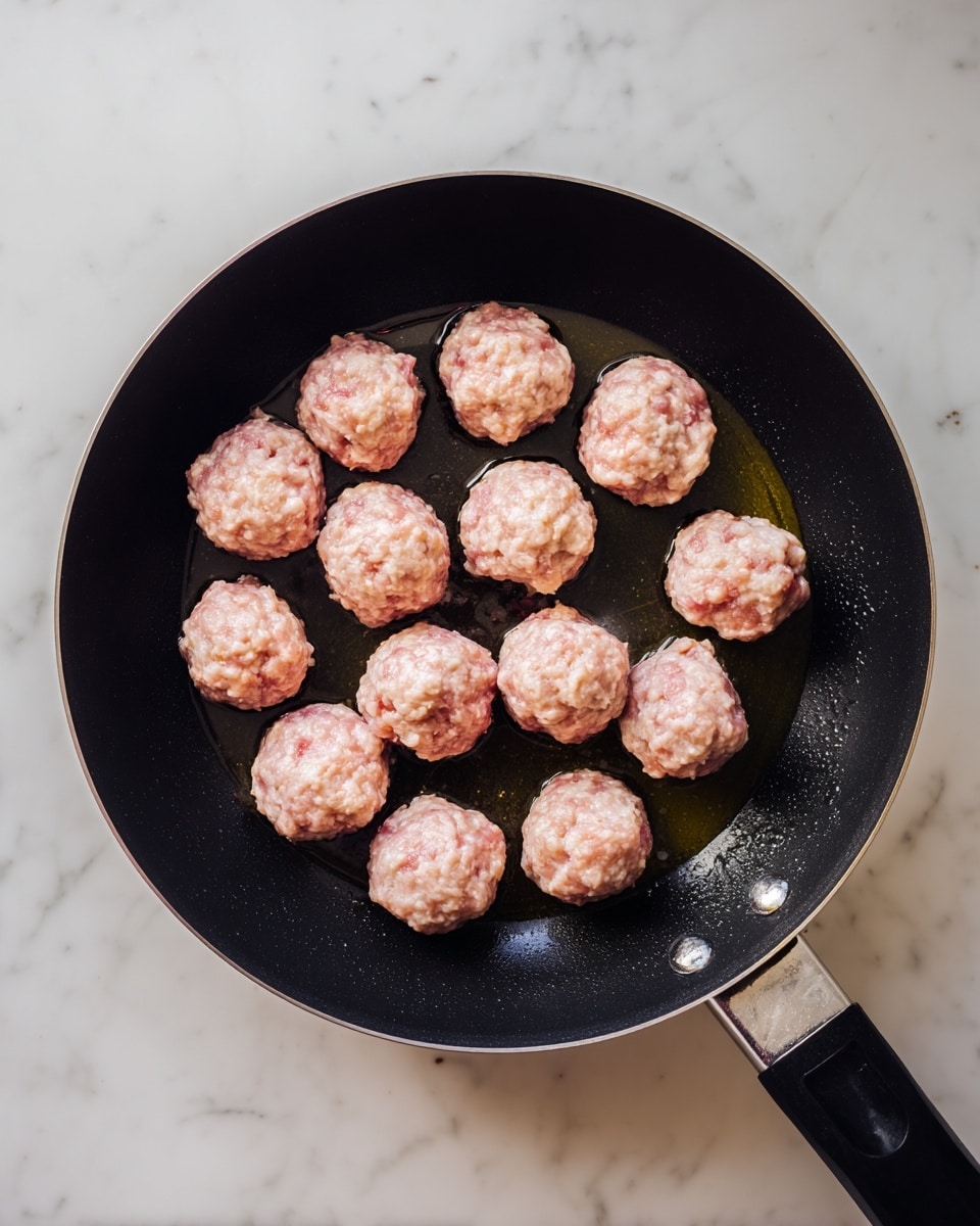 A black frying pan holds twenty small raw meatballs arranged in a loose circular pattern inside the pan. The meatballs are light pink with visible bits of white and beige, showing a coarse texture. The pan has a thin layer of oil shining under the light, and the background is a white marbled surface. Photo taken with an iphone --ar 4:5 --v 7