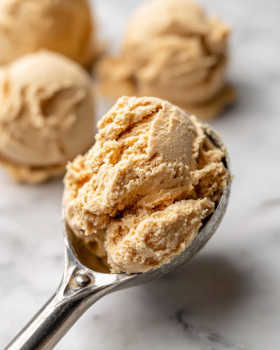The image shows a close-up of a shiny metal ice cream scoop holding a scoop of thick, light brown dough with visible texture and small air pockets. The dough looks soft and sticky, with some parts slightly rough. In the blurred background, there are more scoops of the same dough on a white marbled surface. The focus is sharp on the dough in the scoop, highlighting its texture and color. Photo taken with an iphone --ar 4:5 --v 7
