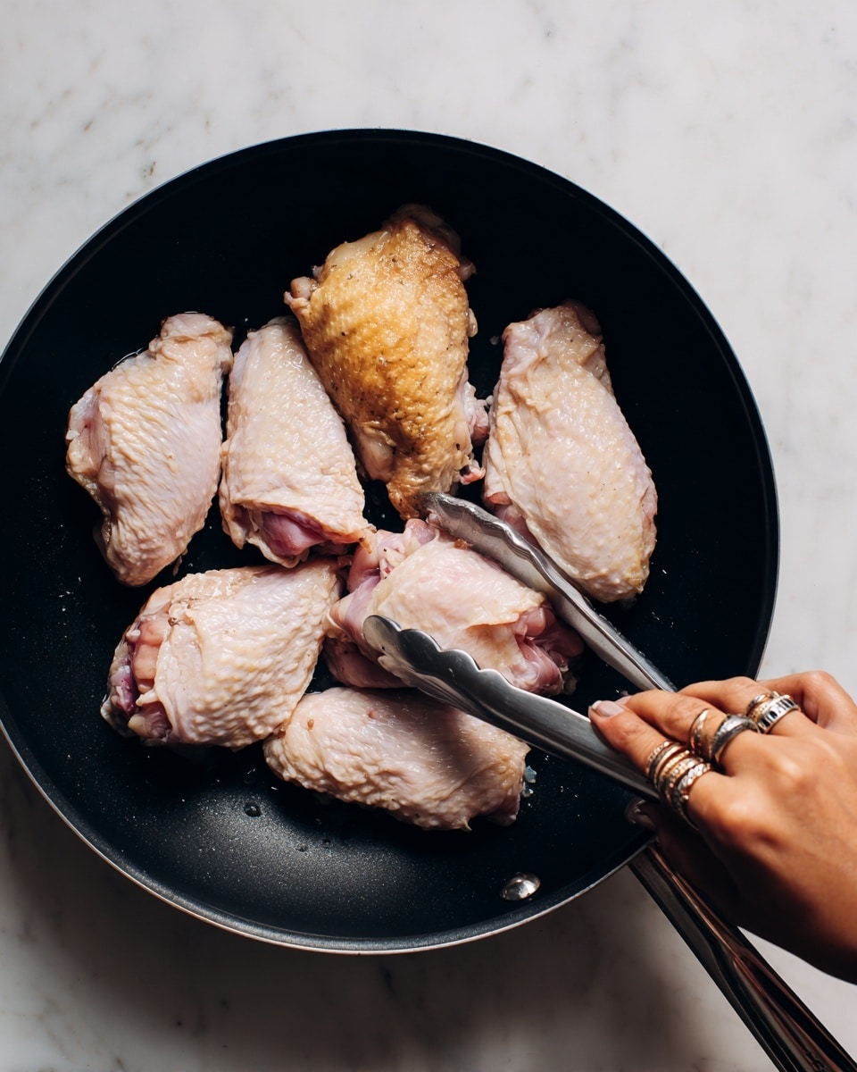 A black frying pan with five pieces of chicken skin side up and raw, light pink chicken skin side down, arranged inside the pan. The chicken skin on one piece is golden and cooked, while the rest show raw pale pink and white textures. A woman's hand with silver rings is using metal tongs to lift or turn one of the raw chicken pieces. The pan is set on a white marbled surface. photo taken with an iphone --ar 4:5 --v 7
