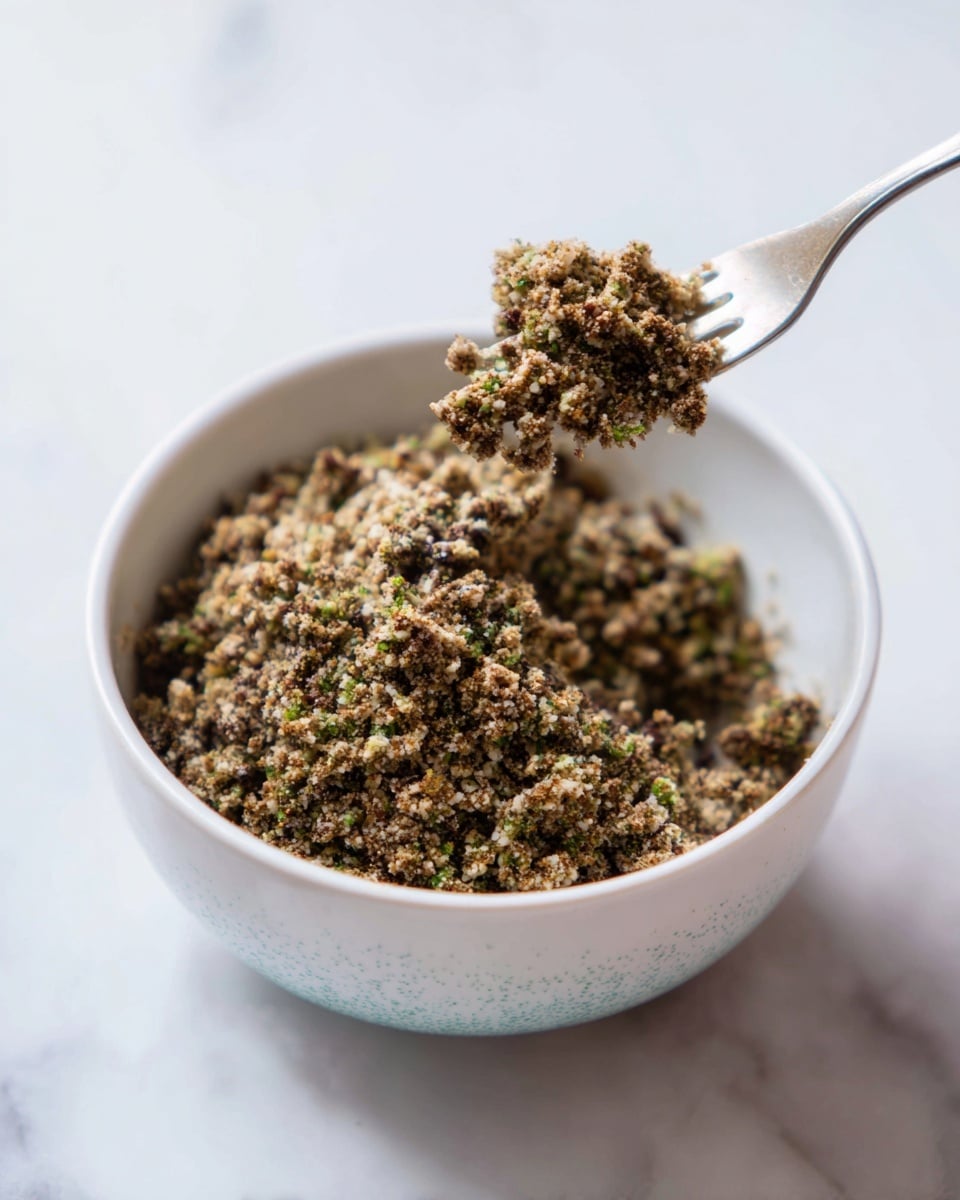 The image shows a close-up of a small, round pastry cut in half and stacked, revealing two layers: a thick golden-brown dough outer layer and a middle filling layer of small green and brown chopped nuts, possibly pistachios, mixed with a crumbly texture. The top of the pastry is sprinkled lightly with fine sugar and tiny purple and white specks. The halves sit on a sheet of light brown parchment paper placed on a white plate with a smooth edge, all set against a white marbled surface. Some small pieces of nuts and red dried flower petals are scattered around the plate. Photo taken with an iphone --ar 4:5 --v 7