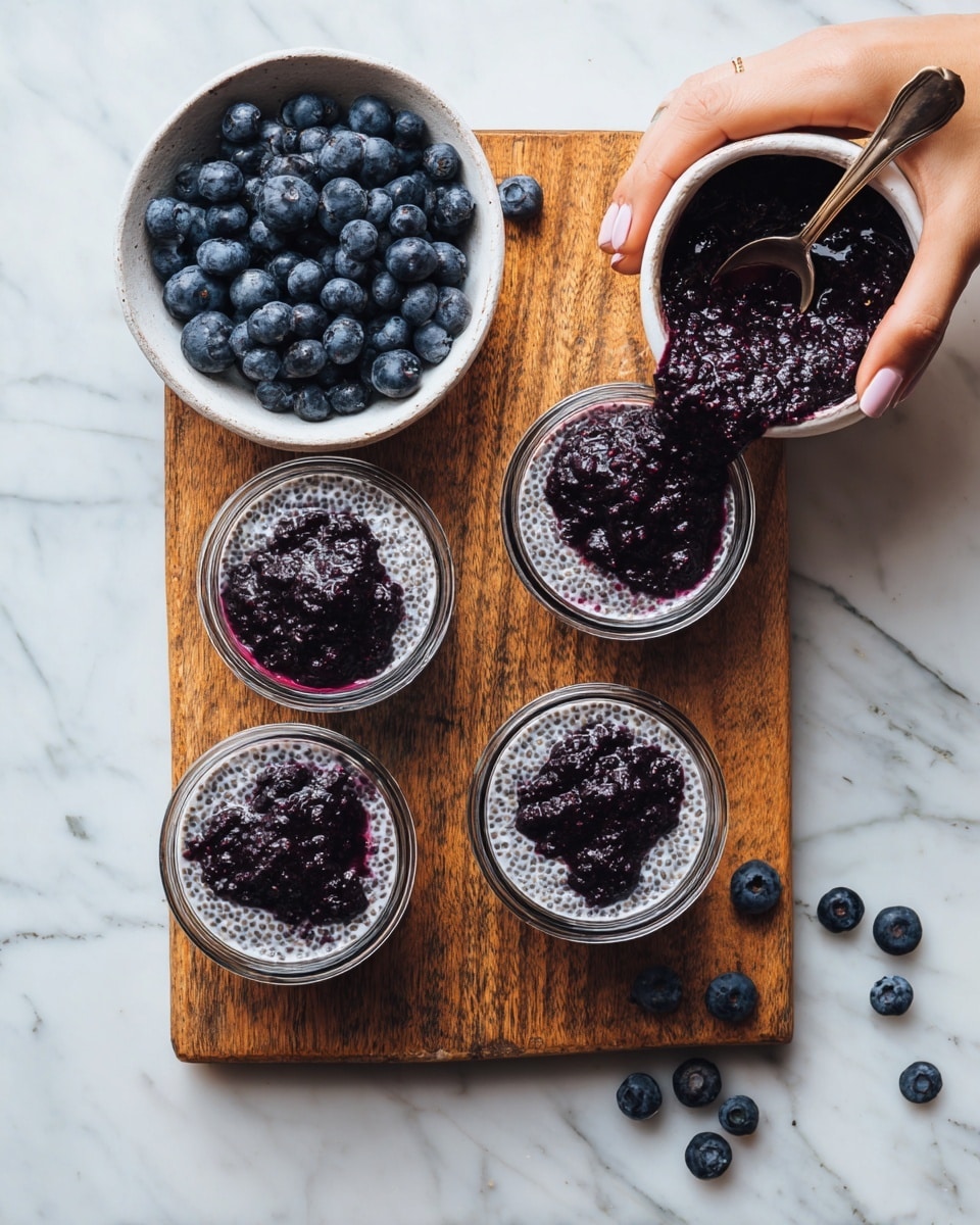 The image shows two parts: on the left side, a white bowl filled with many fresh, dark blue blueberries on a wooden board with a white marbled surface underneath. On the right side, a woman's hand is pouring thick dark purple sauce into one of four clear glass bowls. Each bowl contains a white, textured chia pudding mixed with light gray and white seeds, topped with scattered blueberries. One bowl has visible sauce already poured on the chia pudding with a spoon resting inside. The wooden board and white marbled surface continue here, with some loose blueberries near the bowls. photo taken with an iphone --ar 4:5 --v 7