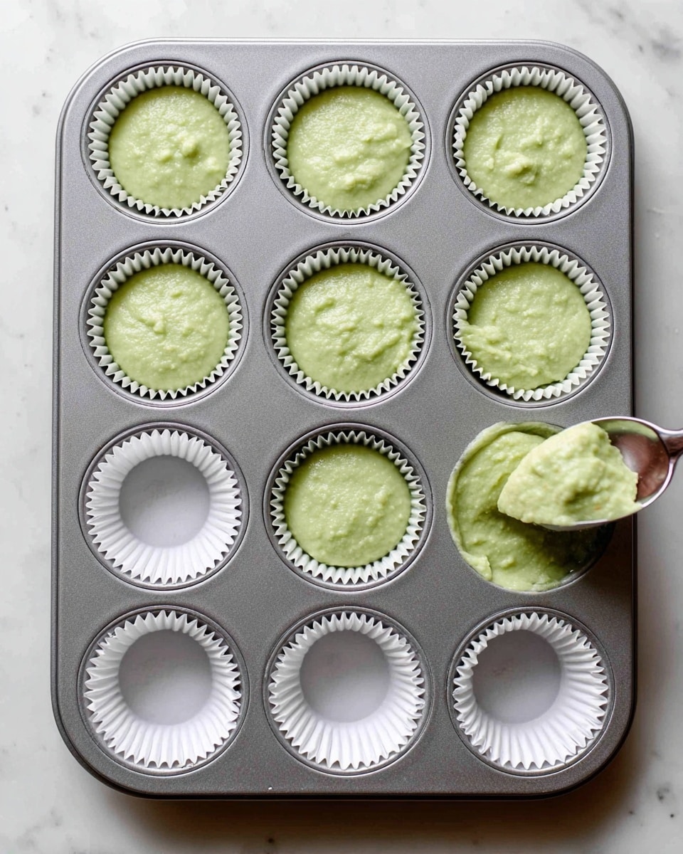 A grey metal muffin tray with twelve spaces, each lined with white paper cups. The top two rows have six cups filled with pale green batter that looks smooth with a few small lumps. The bottom row has six empty white paper cups, with the far right cup being filled using a metal scoop holding the same green batter. The tray sits on a white marbled surface. photo taken with an iphone --ar 4:5 --v 7