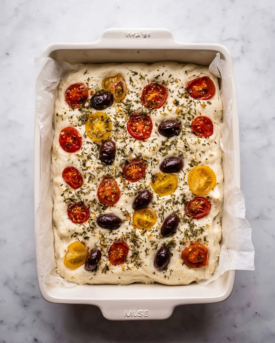 No Knead Focaccia Recipe 5 A white rectangular baking dish lined with parchment paper holds a single thick, fluffy layer of focaccia dough that is pale beige with a soft, spongy texture. On top of the dough are evenly spaced halved red and yellow cherry tomatoes that add bright pops of color, along with dark brown whole olives scattered across the surface. Green dried herbs are sprinkled generously over the entire focaccia, adding texture and contrast. The dish sits on a white marbled surface, and the photo is taken with an iphone --ar 4:5 --v 7