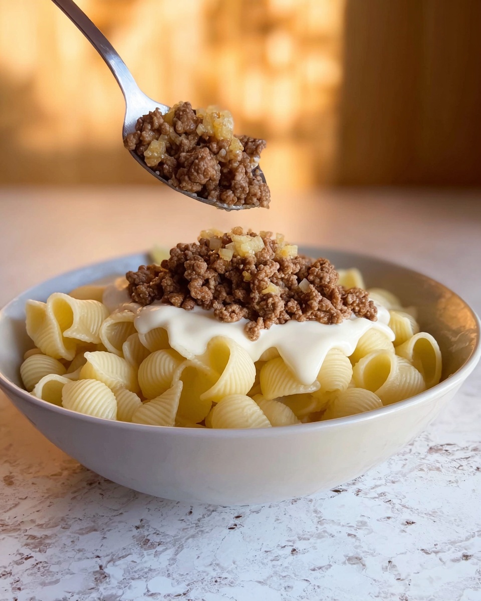 A white bowl holds one layer of small, hollow shell pasta in pale yellow. On top of the pasta is a thick layer of creamy white sauce, and above that is a loose pile of cooked ground meat mixed with diced onions, showing a crumbly, brown texture with some golden bits. A metal spoon is held above the bowl, dropping more of the browned meat mixture slowly onto the layers below. The bowl sits on a white marbled surface with a soft, warm background glow. photo taken with an iphone --ar 4:5 --v 7