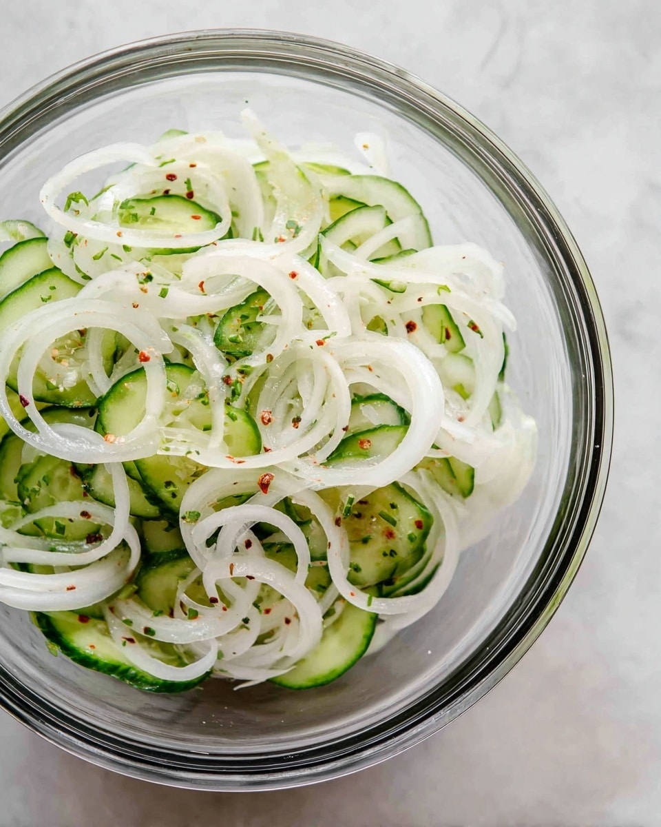 A clear glass bowl holds a mix of thinly sliced white onions and small green cucumber slices, lightly soaked in a clear liquid with tiny red chili flakes sprinkled throughout. The onions form loose curly rings that spread over the cucumber slices, creating a layered look with white and pale green colors on a white marbled surface. photo taken with an iphone --ar 4:5 --v 7
