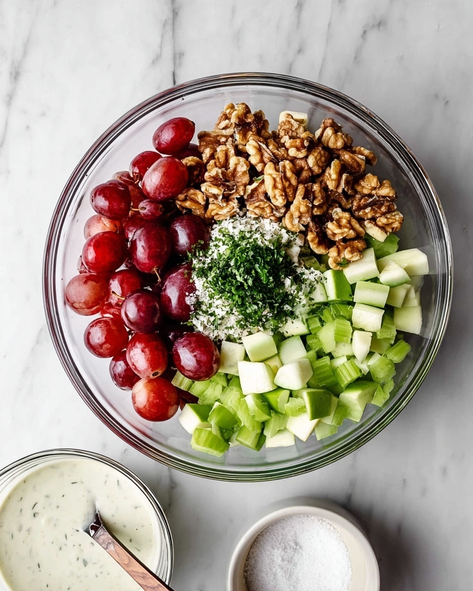 A clear glass bowl on a white marbled surface holds a colorful mix of ingredients arranged in separate sections. There are halved red grapes on the left side, chunky walnut pieces at the top, chopped green celery on the right, and cubed green apple pieces at the bottom. In the middle, a small pile of chopped green herbs and white salt is placed on top of the apple and celery. Below the glass bowl, there is a smaller clear bowl filled with a creamy white dressing with a metal spoon inside, and next to it, a small white bowl with more salt. Photo taken with an iphone --ar 4:5 --v 7