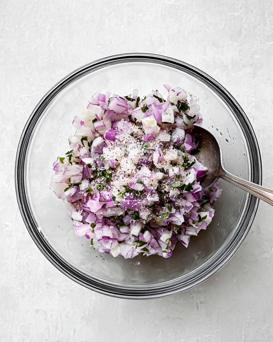 A clear glass bowl sits on a white marbled surface, filled with finely chopped layers of light purple and white onion pieces mixed with small green herb leaves. On top of the onion mix, coarse white salt and black pepper speckles are scattered evenly. A silver spoon rests inside the bowl, partially covered by the chopped mixture. The overall look is fresh, simple, and clean. photo taken with an iphone --ar 4:5 --v 7