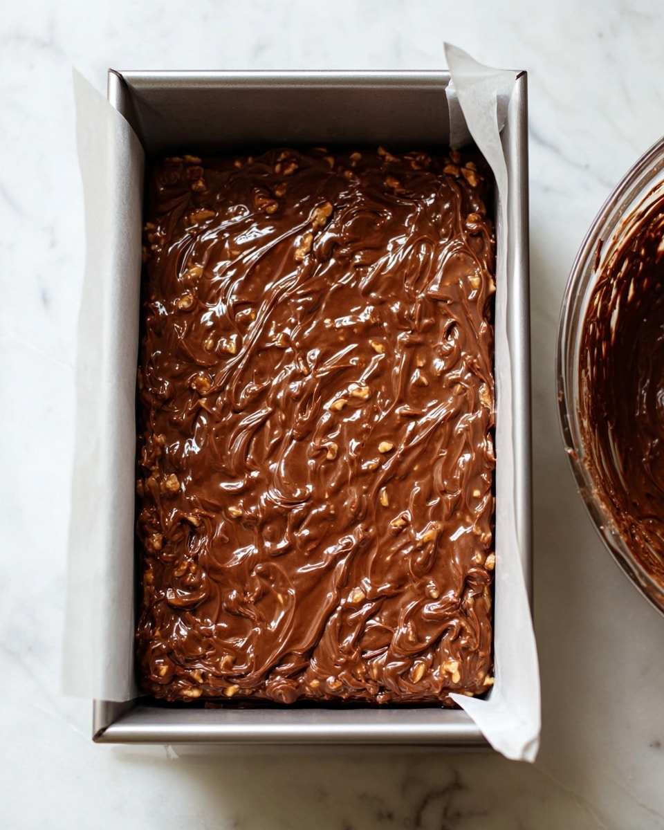 A close-up of a silver rectangular baking pan lined with white parchment paper, filled with one thick layer of chunky chocolate mixture that has a glossy, smooth texture with visible nut pieces mixed in. The pan is placed on a white marbled surface, and to the right side, there is a glimpse of a glass mixing bowl containing leftover chocolate mixture on the sides. The image is bright and clear, showing the uneven, swirled texture of the chocolate layer. Photo taken with an iphone --ar 4:5 --v 7