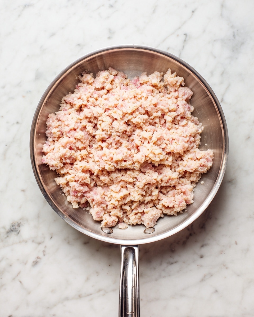 A top view of a silver pan filled with raw ground meat that has a mix of light pink and pale beige colors. The meat is spread evenly inside the pan, showing a rough, crumbly texture. The pan handle is straight and silver, pointing toward the bottom of the image, all placed on a white marbled surface. photo taken with an iphone --ar 4:5 --v 7