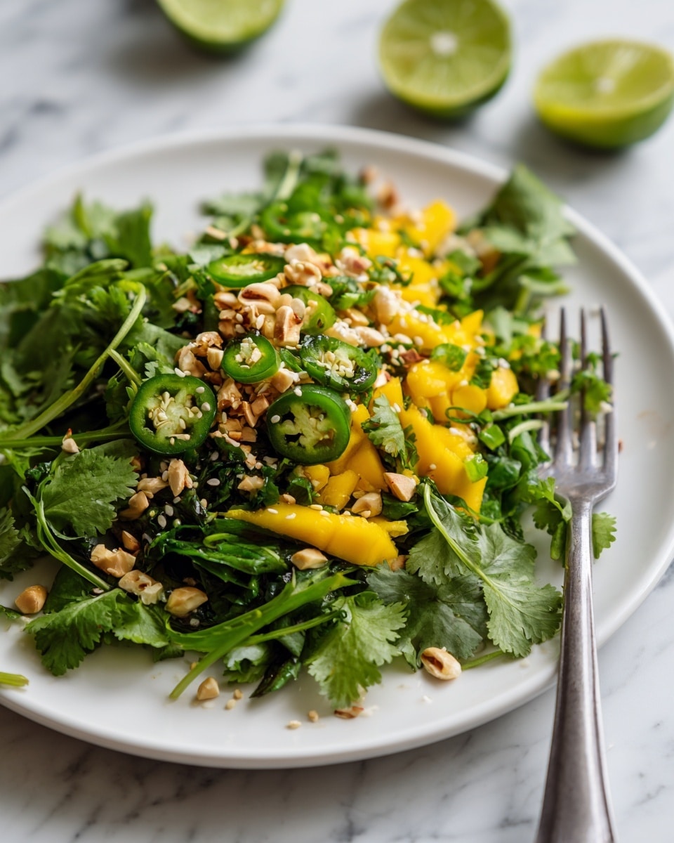 A close-up view of a white plate filled with a colorful salad arranged in layers. The bottom layer is fresh, dark green cilantro leaves, spread across the plate. On the right side, there is a layer of bright yellow mango chunks mixed with small green herb pieces. Layered diagonally across the plate are thin slices of bright green jalapeños and scallions adding texture and freshness. On top of the jalapeños and cilantro, there is a scattering of chopped light brown nuts and white sesame seeds adding crunch. The plate is placed on a white marbled surface, and there are blurred lime halves in the background, hinting at fresh citrus. A silver fork rests on the right side of the plate. Photo taken with an iphone --ar 4:5 --v 7