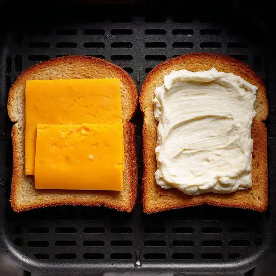 The image shows a sandwich being made on a black air fryer tray with two slices of light tan bread. The slice on the left has one thick, bright orange slice of cheddar cheese placed flat on top. The slice on the right is spread with a thick layer of white mayonnaise on the surface, without any cheese or other ingredients visible. The tray and background have a dark tone, but the air fryer mesh texture is clear under the bread. photo taken with an iphone --ar 4:5 --v 7