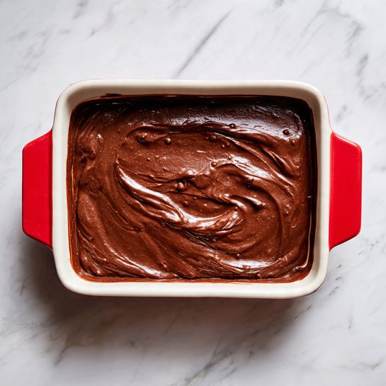 A close-up top view of a rectangular dish filled with one thick layer of smooth, dark brown chocolate batter evenly spread inside. The batter has a slightly shiny, sticky texture with some gentle swirls and small air bubbles visible on the surface. The dish is white inside with a red border around the edges, placed on a white marbled surface. photo taken with an iphone --ar 4:5 --v 7